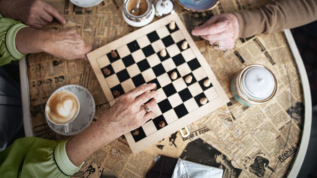 A bird's eye view of three older adults having coffee and playing chess on a table. 