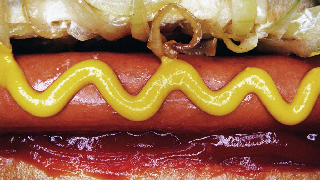 A close-up of a hotdog with mustard, tomato ketchup, and onions, depicting ultra-processed foods